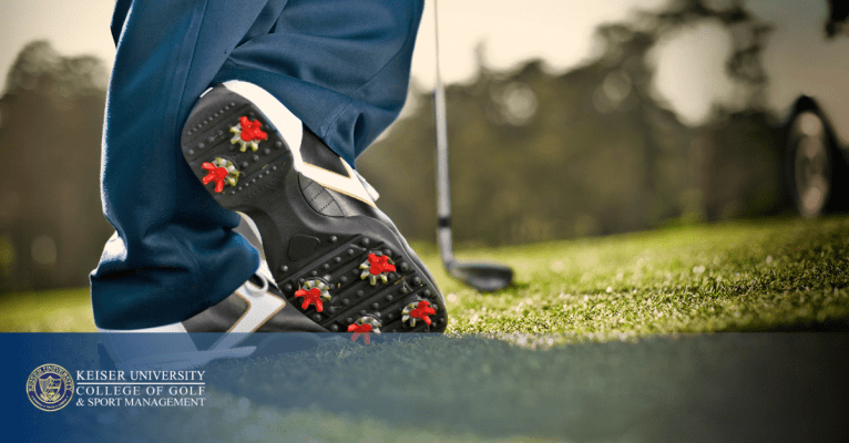 Close-up of spiked golf shoe and club on grass, branded with Keiser University College of Golf logo.