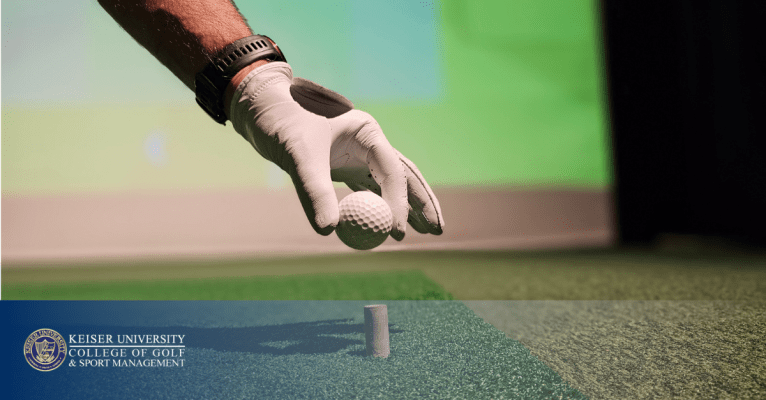 Gloved hand placing a golf ball on tee in indoor simulator, branded with Keiser University College of Golf logo.